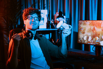 Fototapeta premium Host channel of young gaming streamer looking at camera, holding joystick with fighting Moba battle arena game with multiplays team against on pc screen background at neon digital light room. Gusher.