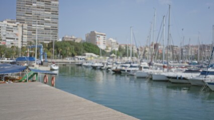 Outdoor waterfront scene with blurred buildings and marina featuring boats and yachts docked along the pier against a clear sky and distant urban backdrop.