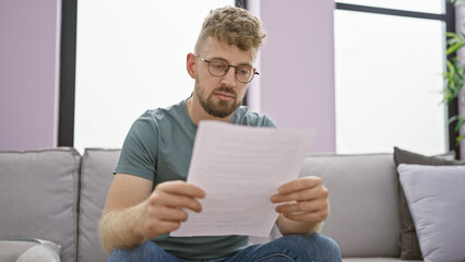 Caucasian man with beard and blue eyes reading a document in a well-lit living room on a grey sofa.