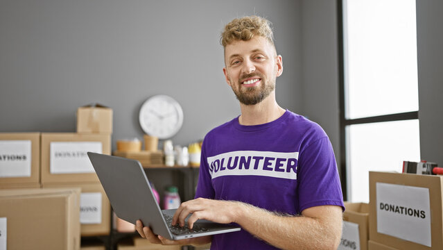A cheerful caucasian man with a beard volunteering at a donation center, holding a laptop in a room full of boxes. - Powered by Adobe