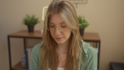 A contemplative blonde woman with glasses in a green sweater indoors, embodying serenity and domesticity.