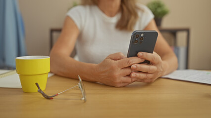 Middle-aged woman using smartphone at home, with coffee cup, glasses, and notebook on table.