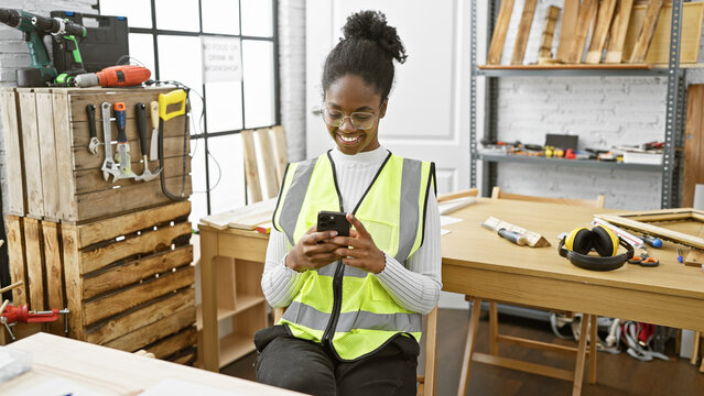 Smiling african american woman in safety vest using smartphone in a well-equipped carpentry workshop