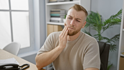 A young caucasian man with a beard feeling a toothache in a modern office setting