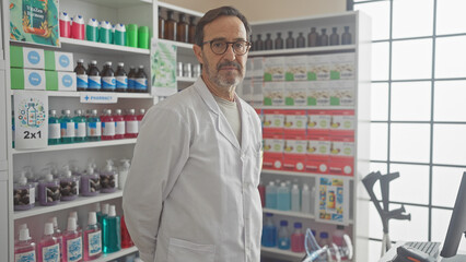 A mature hispanic man in a white coat stands confidently inside a pharmacy full of medical supplies.