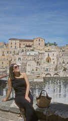 Fototapeta premium A young hispanic woman in sunglasses sitting at a viewpoint in the old town of matera, basilicata, italy, enjoying the sunny weather against a backdrop of ancient buildings and blue sky.