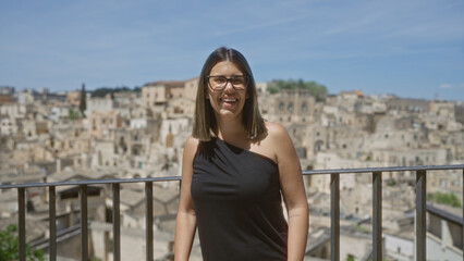 Fototapeta premium A young hispanic woman stands at a viewpoint in the old town of matera, basilicata, italy, with ancient stone buildings in the background under a clear blue sky, smiling beautifully.