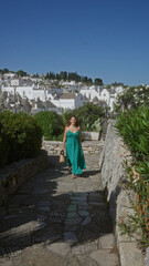 A young hispanic woman walks along a charming street in the old town of alberobello, puglia, italy, surrounded by trulli houses under a clear blue sky.