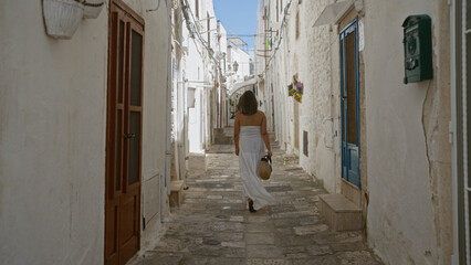 A young hispanic woman strolls through the charming old town of ostuni in puglia, italy, wearing a white dress and carrying a wicker basket.