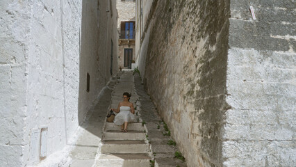 Young hispanic woman in a white dress seated on narrow stone steps in the historic old town of ostuni, puglia, italy, surrounded by tall whitewashed buildings on a bright sunny day.