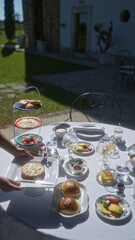 A woman's hand places a plate on a well-decorated breakfast table outdoors at an italian villa in puglia, europe, showcasing an elegant spread of pastries and fruits in the morning sunlight.