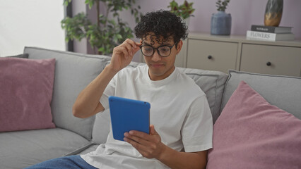 Handsome young hispanic man in glasses using tablet on couch indoors