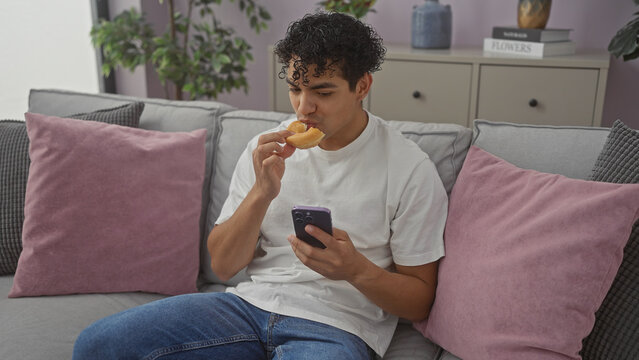 A handsome young hispanic man in casual clothing eats a snack while using a smartphone on the couch in a modern living room setting.