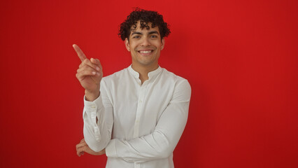 A cheerful young hispanic man in a white shirt pointing up, standing against a vibrant red isolated background.