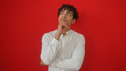 Pensive young hispanic man in white shirt over isolated red background contemplating