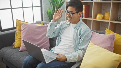 Happy asian man waving while video calling from his comfortable living room couch.