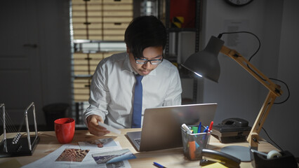 A young asian man analyzes documents on a laptop in a dimly lit indoor office at night.
