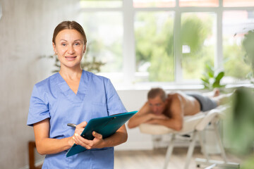 Female massage therapist wearing medical uniform standing at sunny massage room holding folder in her hands