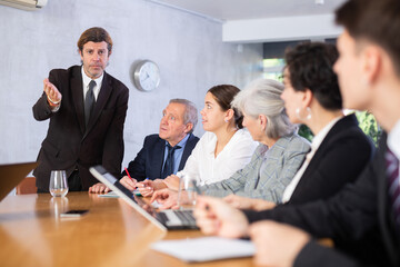 Portrait of interested adult entrepreneur in formal suit explaining new business strategy to partners sitting at table in office