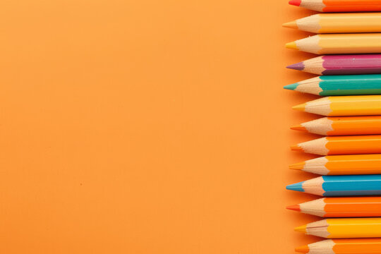A row of colorful pencils lined up on an orange background, creating space for text or graphics. The pencils have different colors and shades to create contrast and visual interest. 
