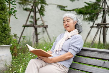 A calm mature Asian woman is sitting on a bench, her eyes closed and deep in thought while reading.