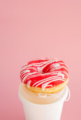 Donut with pink icing and white stripes rests on takeaway coffee cup.