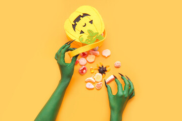 Green hands of witch with bag and tasty candies on orange background. Halloween celebration