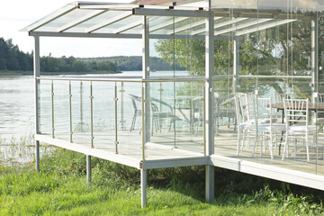 Glass modern gazebo with table and chairs near lake. Buildings, structures in nature in summer. 