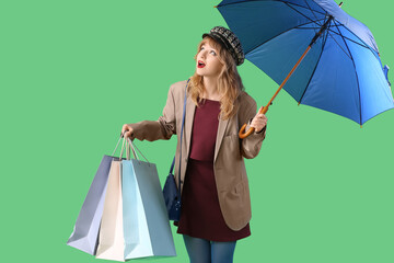 Stylish young woman in fall clothes with umbrella and shopping bags on green background