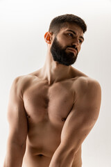 Fototapeta premium Shirtless young man with beard and short hair looks pensively to side, showing off his muscular physique against white background. Concept of male health, body care, natural beauty people, fitness.
