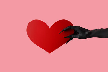 Black hand of witch with paper heart on pink background. Halloween celebration