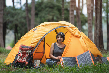 Asian female backpacker sitting at her tent at campsite in pine forest, portable solar panel place nearby, looking at at camera, journey with backpack concept