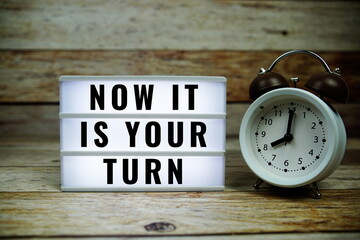 Now it is your turn text on LED lightbox with alarm clock on wooden background, Business and Money...