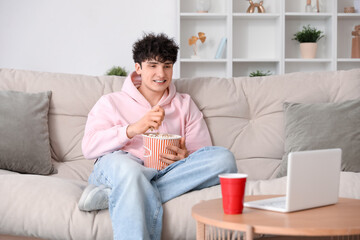 Obraz premium Young man watching movie with laptop and eating popcorn on sofa in living room