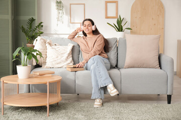 Beautiful young woman in headphones listening to audiobook with books and sitting on sofa in living room