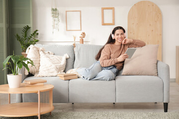 Obraz premium Beautiful young woman in headphones listening to audiobook with modern tablet computer and sitting on sofa in living room