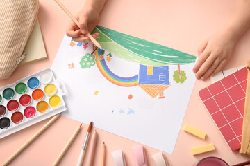 Child's hand drawing rainbow with pencils, watercolor paints and different stationery on beige...