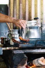 A close-up view of a sausage being grilled on a rustic stove, with a hand using tongs and metal...
