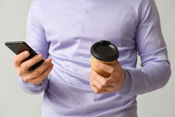 Young businessman with paper cup of coffee and mobile phone on grey background, closeup