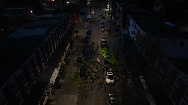 Nighttime view of a residential street in American historic neighborhood. Row homes and parked cars. Dimly lit streets. Aerial reverse dolly.