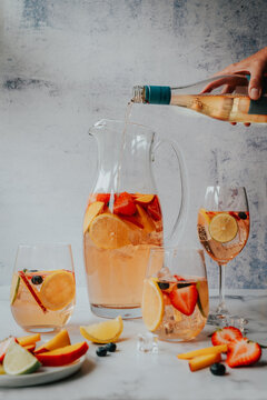 Hand pouring wine into pitcher of white sangria beside filled glasses.