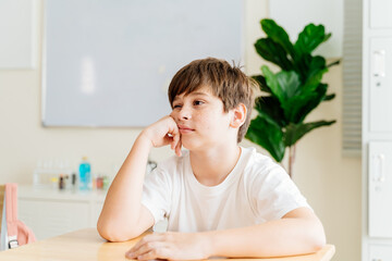 Fototapeta premium Freckled boy in classroom setting, thinking and daydreaming. Young student in white shirt at desk, casual and relaxed, showcasing youth and innocence. Education and learning environment portrait