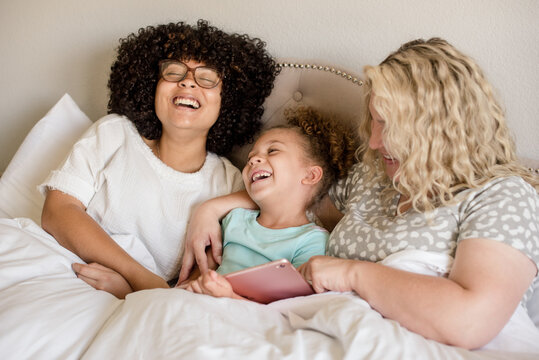 Mother and daughters are laughing while holding a tablet in bed - Powered by Adobe