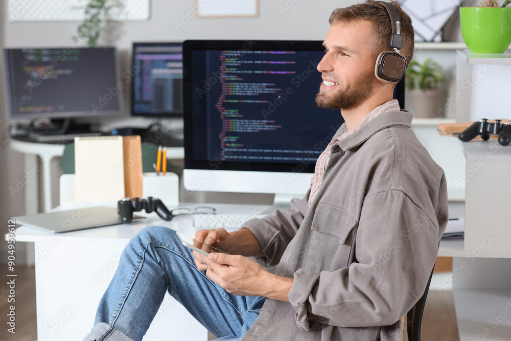 Wall mural young bearded programmer with headphones and tablet computer working at table in office