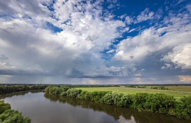 A tranquil river meanders through a vibrant green landscape under a dramatic sky filled with dynamic clouds during the golden hour.