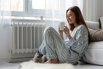 Young woman wearing a sweater freezing for winter cold with her cat. Girl is sitting over electric heater, drinking hot coffee or tea, warming up hands. Discomfort spending time at home. Coldly, chill