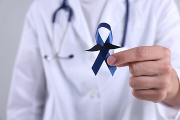 Prostate cancer awareness. Doctor holding blue ribbon with fake mustache on light grey background,...