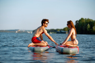 Young couple spending sunny day on lake, paddle boarding. Muscular man in sunglasses helping with with her board. Concept of sport, summer, active lifestyle, hobby, leisure time