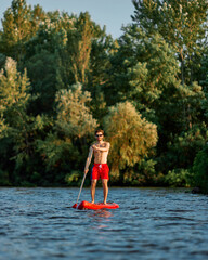 Young man in red swim shorts, with muscular body standing on paddle board in the middle of lake and...