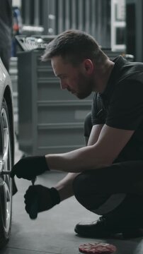 Medium vertical shot of Caucasian tire fitter walking up to white clients car, crouching down, tightening bolts on wheel with wrench after maintenance in garage